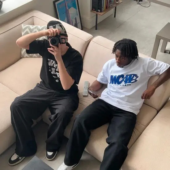 Two people sitting on a couch wearing fashionable clothing, one is looking through a camera.